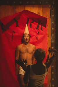ORAISON spectacle cirque audiodescription - photo d'un homme torse nu sur un fond rouge face à une lançeuse de couteaux
