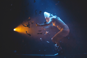ORAISON spectacle cirque audiodescription - photo d'un homme torse nu sur un fond bleuté avec une lumière jaune, un comédien en équilibre dans une ambiance feutrée 
