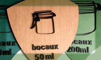 EMERAUDE ID signalétique accessible indiquant le rangement des bocaux par 50m, 100ml et 200 ml. Panneau en forme de goutte avec la pointe vers le bas. Le visuel du bocal et le texte sont en relief en bois noir sur fond coloré.