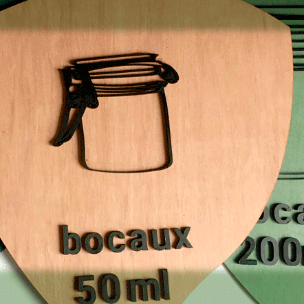 EMERAUDE ID signalétique accessible indiquant le rangement des bocaux par 50m, 100ml et 200 ml. Panneau en forme de goutte avec la pointe vers le bas. Le visuel du bocal et le texte sont en relief en bois noir sur fond coloré.