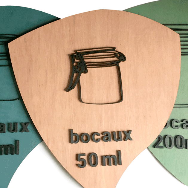 EMERAUDE ID signalétique indiquant le rangement des bocaux par 50m, 100ml et 200 ml. Panneau en forme de goutte avec la pointe vers le bas. Le visuel du bocal et le texte sont en relief en bois noir sur fond coloré.