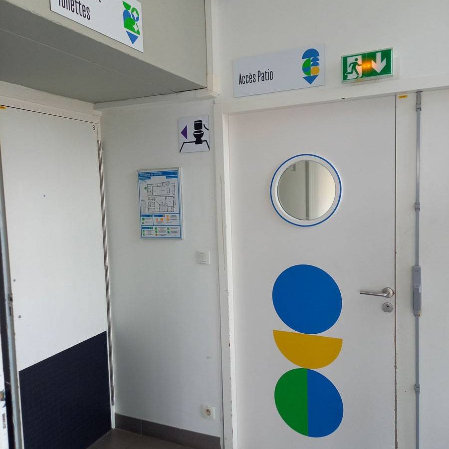 La signalétique de Bonjour Minuit a été pensée en 3 zones. Chaque zone est identifiée par une dominante de couleur et une dominante de forme. Ici nous voyons la pièce par laquelle les personnes rentrent le jour. Elle est identifiée par des ronds et demi cercles en majorité bleus et verts. Ces formes se retrouvent aussi bien dans le décor des portes quand sur les panneaux directionnels